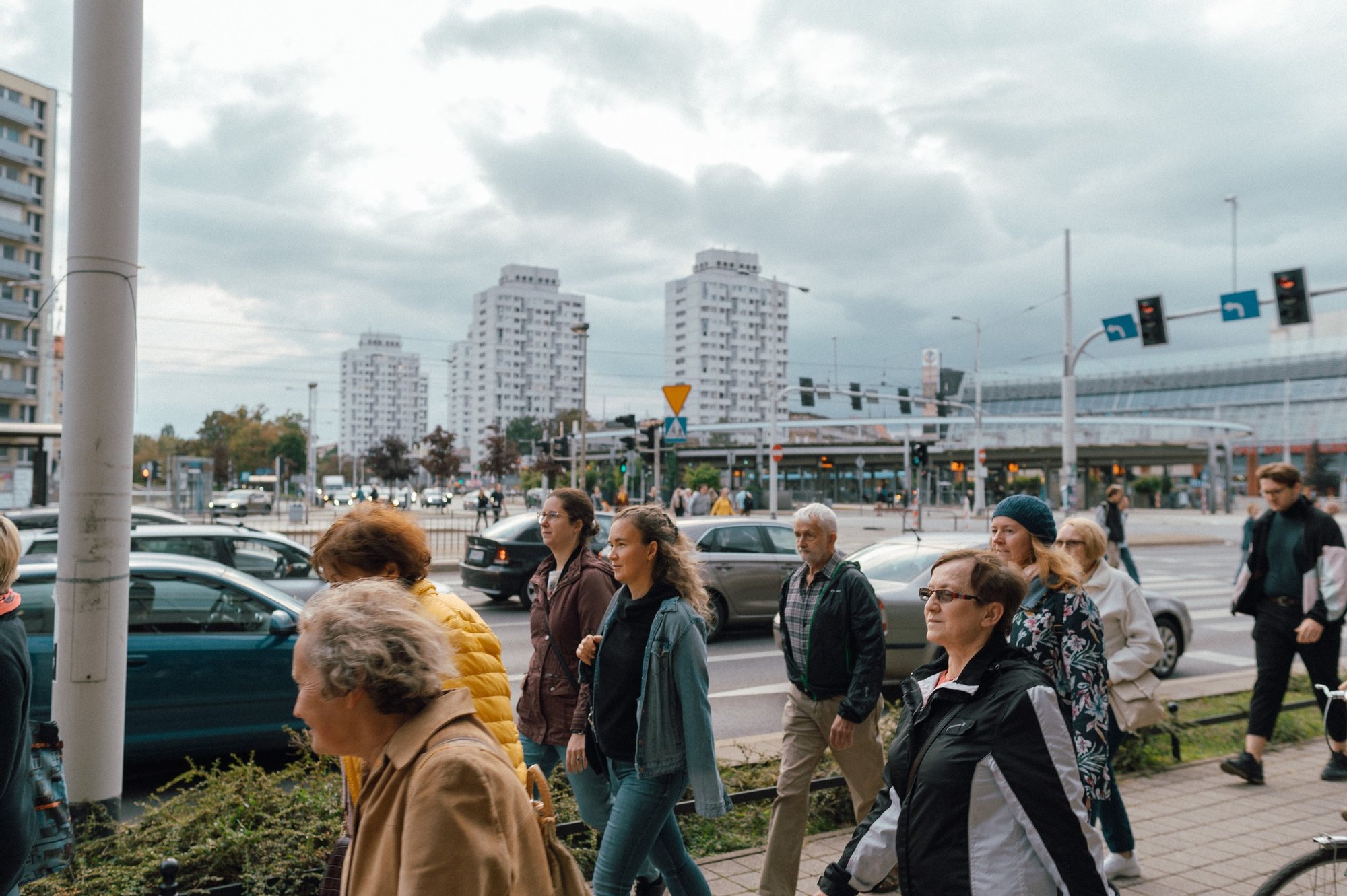 Spacerująca grupa osób w różnym wieku. W tle rondo Reagana i górujące nad nim Sedesowce.