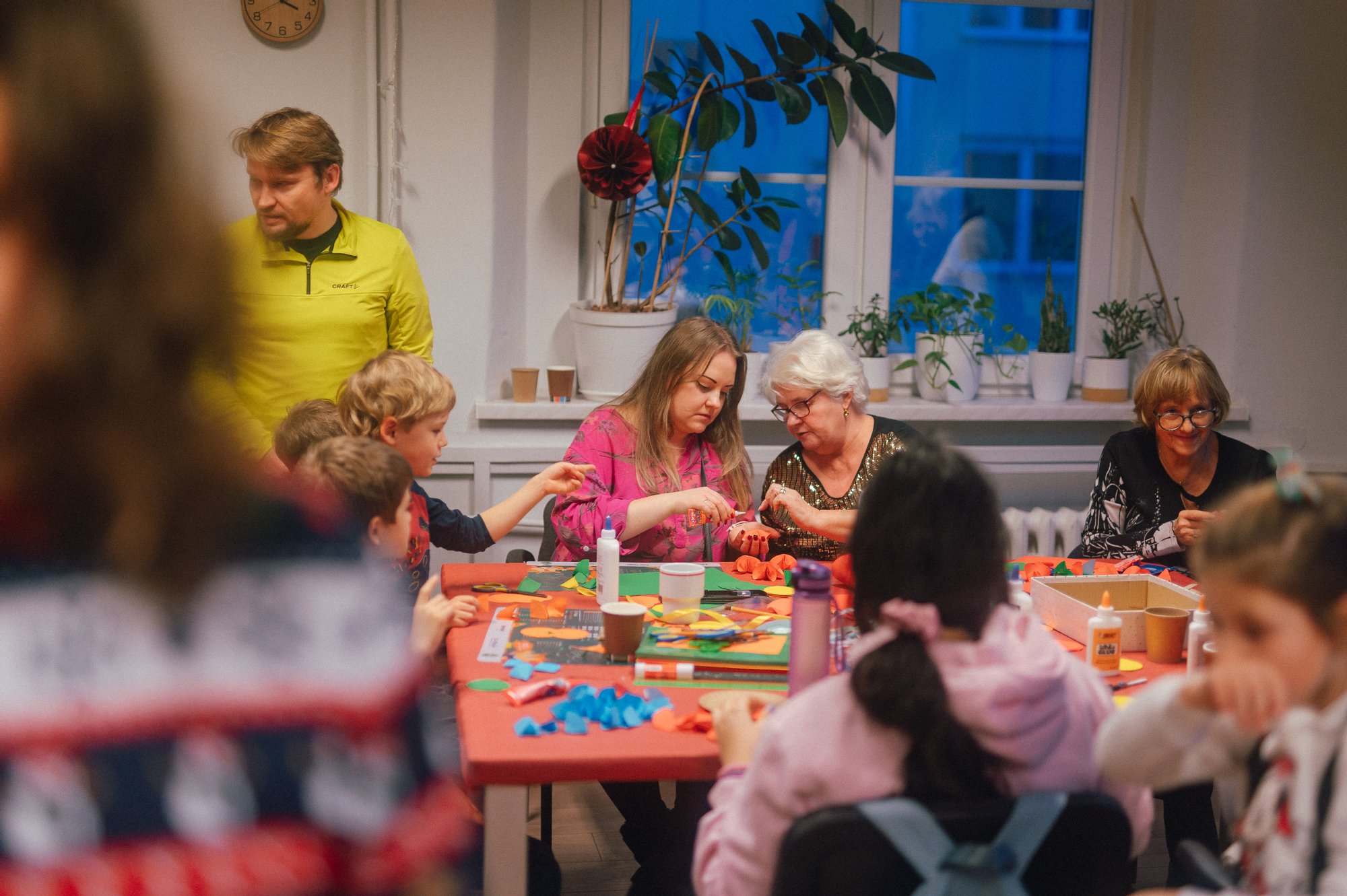 Kolorowa, pozioma fotografia: w jasnym pomieszczeniu przy dużym stole siedzi grupa osób w różnym wieku, zarówno dzieci, jak i seniorek. Starsza kobieta razem z młodą dziewczyną kleją razem ozdoby choinkowe. Na stole porozkładanych jest dużo materiałów plastycznych. Dookoła stołu kręcą się inni ludzie. Trwa sąsiedzkie spotkanie świąteczne.