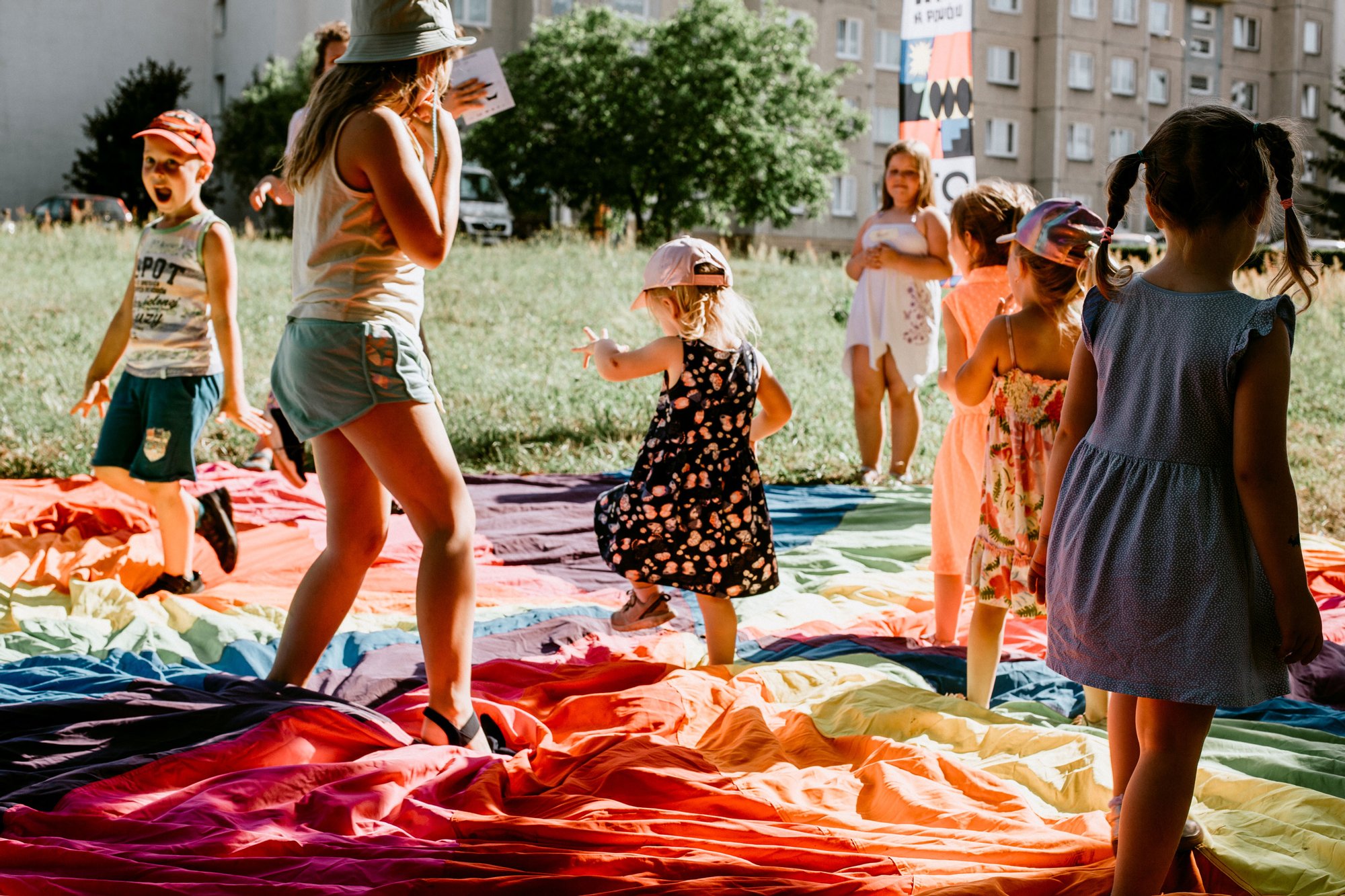Na trawie między blokami rozłożona jest kolorowa chusta, po której biegają dzieci w różnym wieku. Jest gorąco i słonecznie.