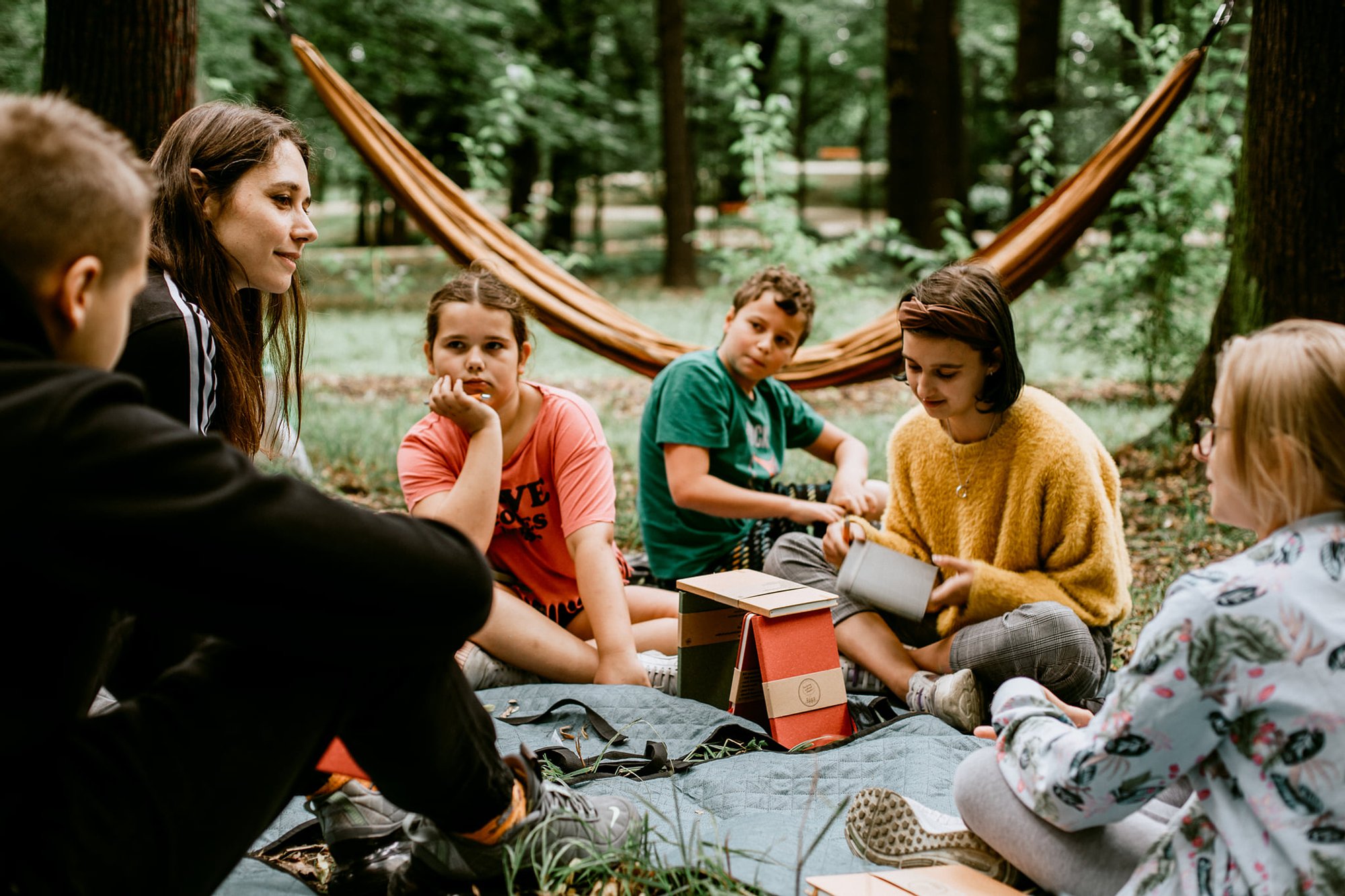 fot. Kalina Żaczek Grupa dzieciaków siedzi na trawie między drzewami, za nimi rozwieszone są hamaki. Trwają warsztaty.