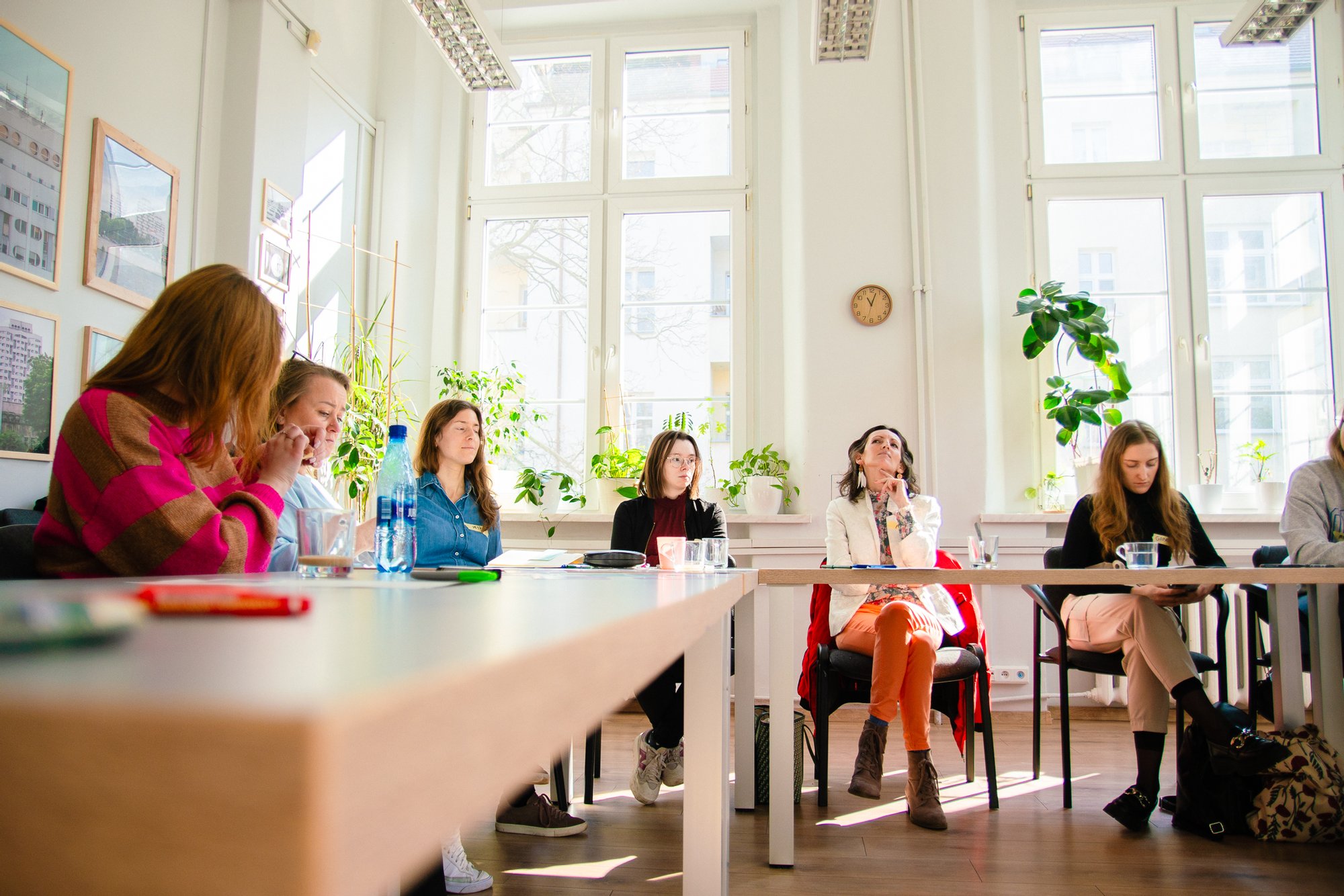Kolorowa fotografia zrobiona w trakcie warsztatów. Przy długich stołach w jasnym pomieszczeniu siedzi grupa kobiet w różnym wieku.