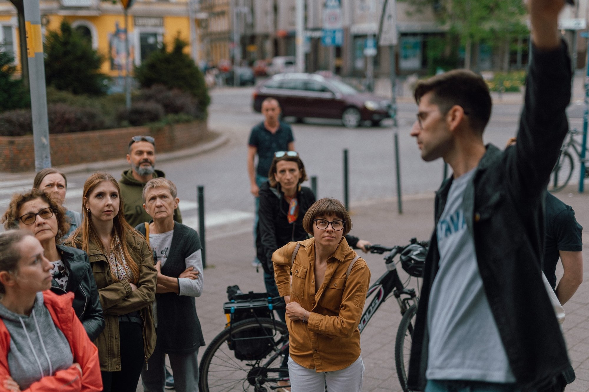 fot. Marcin Szczygieł Kolorowa, pozioma fotografia przedstawia grupę ludzi podczas osiedlowego spaceru. Przewodnik w okularach wskazuje na ścianę budynku.