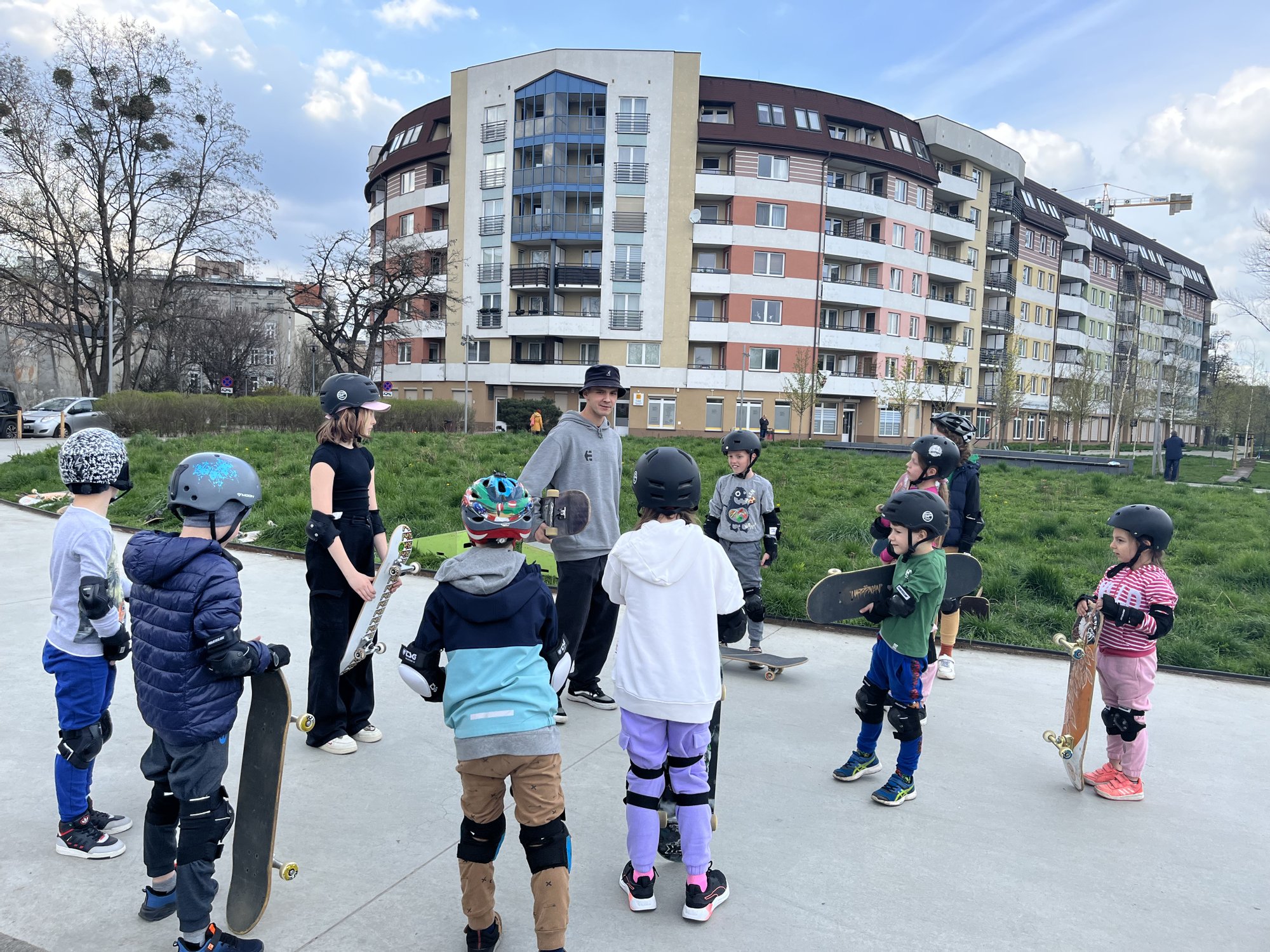 Kolorowa, pozioma fotografia. Dziesiątka dzieci zebrała się na betonowym chodniku. Każde z nich ma kask i ochraniacze, a w ręce trzyma deskorolkę. Dzieci zebrały się wokół młodego mężczyzny, który poprowadzi warsztaty.