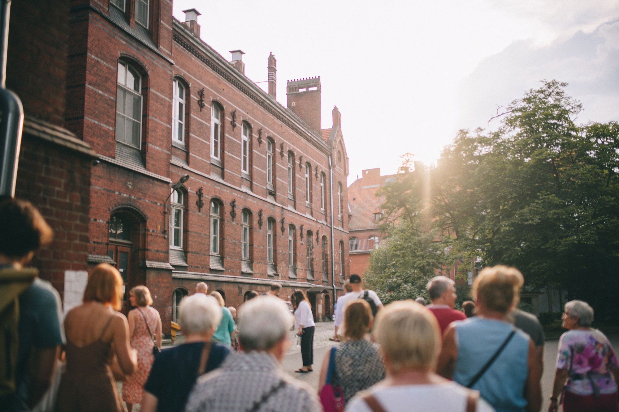 Kolorowa fotografia przedstawia grupę osób spacerujących po osiedlu plac Grunwaldzki. Ostrość zdjęcia uchwycona jest na stare ceglane kamienice.