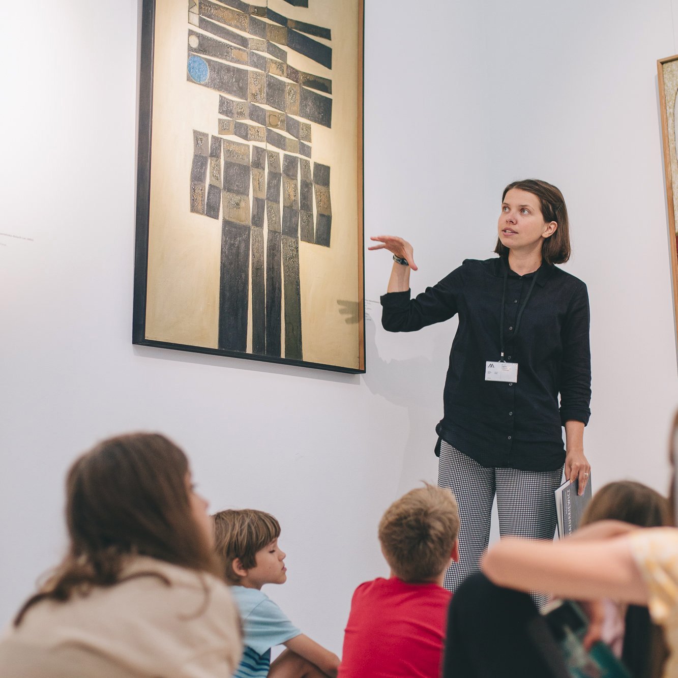 zdjęcie: Marta Sobala Kolorowa fotografia wykonana podczas wycieczki do muzeum. Młoda kobieta stoi przy obrazie i opowiada o nim, na podłodze siedzą zasłuchane dzieci.