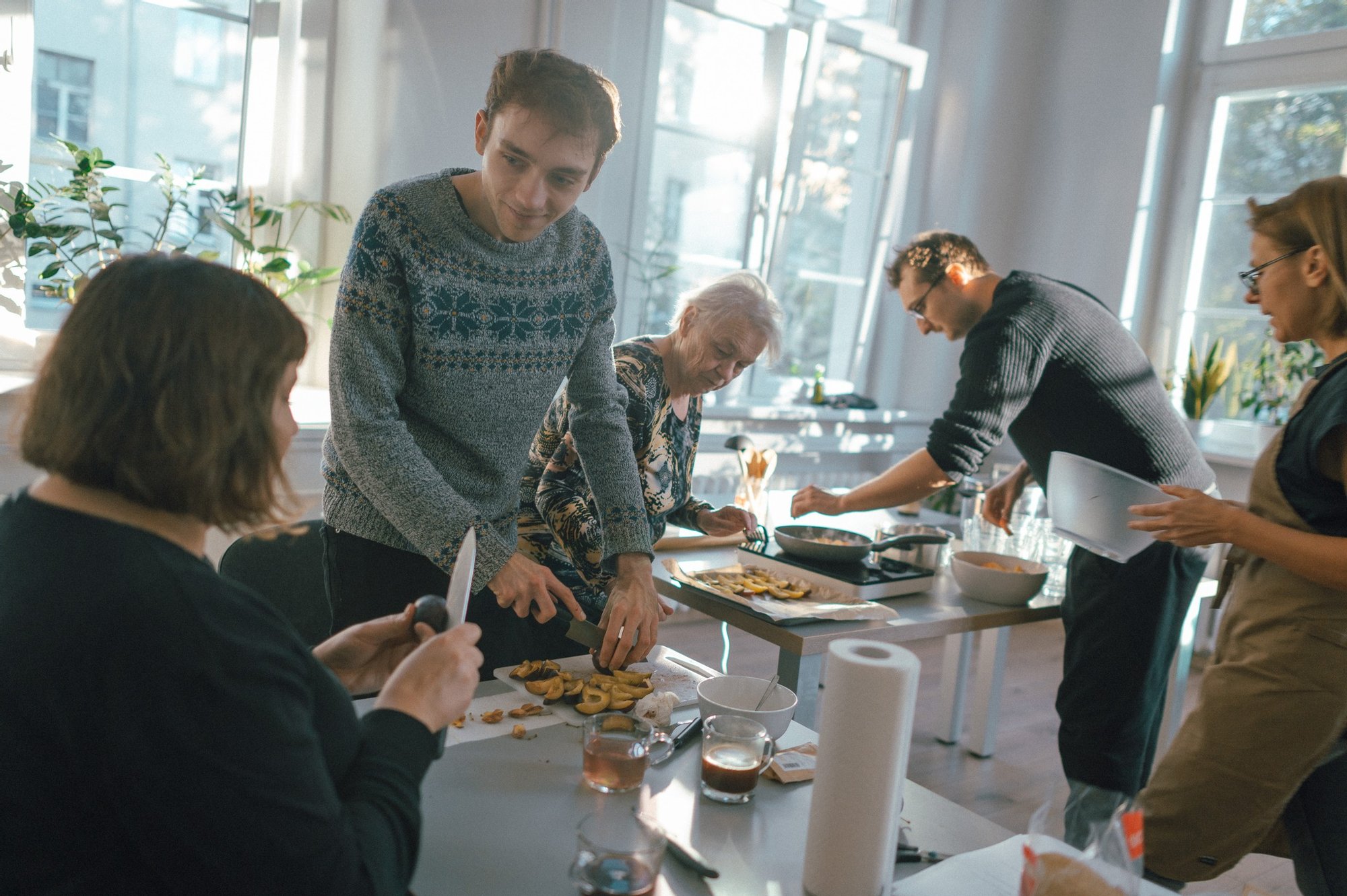 Kolorowa fotografia wykonana podczas warsztatów kulinarnych, grupa ośób w różnym wieku przygotowuje potrawy. Dwie osoby kroją owoce, kobieta na drugim planie doprawia przygotowane jedzenie, mężczyzna obok niej smaży coś na patelni.