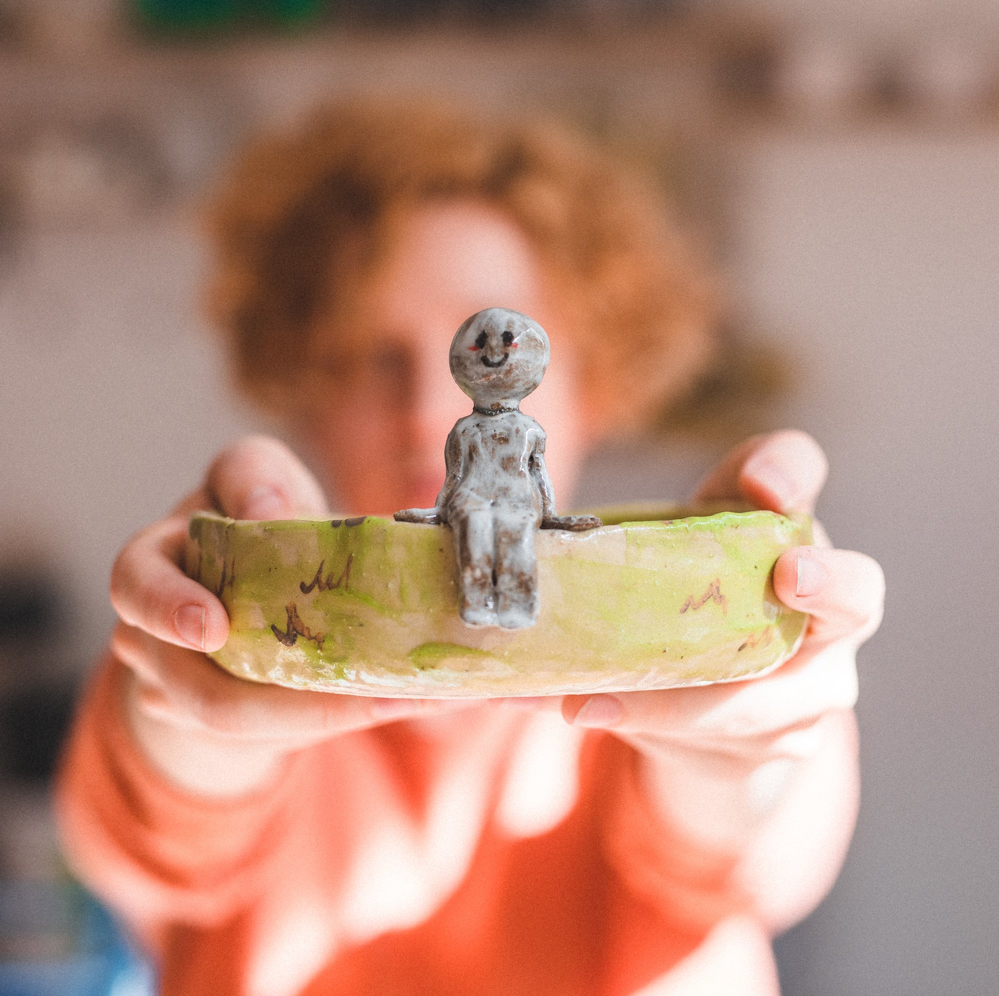 Młoda kobieta wyciąga ręce w kierunku aparatu, trzyma w nich glinianą miseczkę, na brzegu której siedzi uśmiechnięty człowieczek. Ostrość zdjęcia jest na miseczce, osoba z tyłu jest niewyraźna.